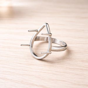 May include: A silver-colored ring with a teardrop-shaped setting. The setting has four prongs extending outwards. The ring is on a light-colored wooden surface.