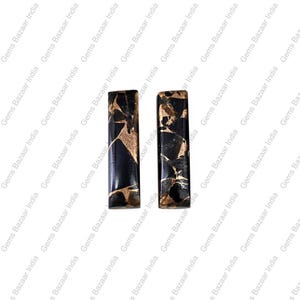 Puede incluir: Dos piedras preciosas rectangulares y pulidas. Las piedras son negras con intrincados patrones de vetas doradas. Las piedras preciosas se muestran sobre un fondo blanco, mostrando sus colores y texturas contrastantes. Probablemente se utilizan para la fabricación de joyas.