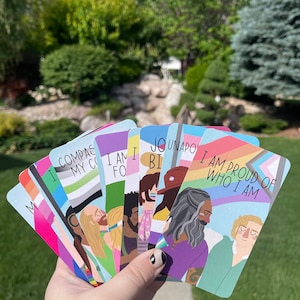 May include: A hand holding a fan of colorful cards with illustrations of diverse people and pride flags. Each card has a different message, including "I am proud of who I am" and "I am enough". The background is a green lawn and trees.