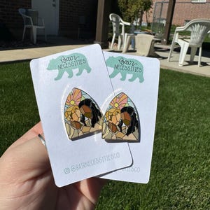Puede incluir: Dos pines de esmalte con un diseño de estilo vitral de dos personas abrazándose. Los pines están en tarjetas blancas con el texto "Bair Necessities" y el nombre de usuario de las redes sociales "@bairnecessitiesco".