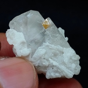 Puede incluir: Un cristal transparente y brillante con un pequeño cristal dorado en forma de pirámide en la parte superior. El cristal está sentado sobre un lecho de material blanco y pulverulento.