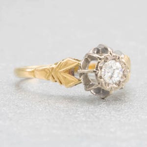 May include: A vintage engagement ring with a gold band and a floral-shaped setting. The setting holds a round, clear diamond. The ring's design features intricate details and a classic aesthetic, suitable for a special occasion.
