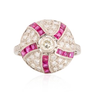 May include: A round, silver ring with a central diamond surrounded by smaller diamonds and rectangular pink gemstones. The ring's design features a radial pattern of pink gemstones and diamonds, creating a striking visual effect. The ring is set against a white background.