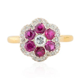 Peut inclure: Bague en or avec un motif floral. Elle présente un diamant clair central entouré de six rubis rouges et de petits diamants clairs. La monture est en métal argenté et la bande est en or.