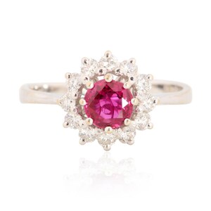 May include: A silver ring featuring a large, round, vibrant pink gemstone at the center, surrounded by a halo of smaller, clear gemstones. The ring's band is smooth and polished, and the setting creates a floral or starburst design.