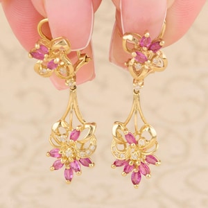 May include: A pair of gold-colored drop earrings. Each earring features a floral design with pink gemstones and small white gemstones. The earrings have a delicate, elegant appearance, suitable for special occasions.