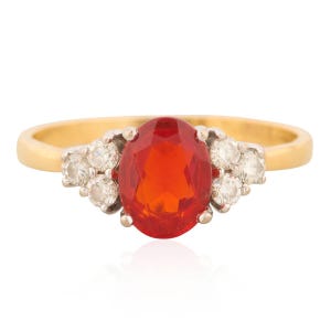 May include: A gold ring featuring an oval, fiery orange gemstone as the centerpiece. Flanking the central stone are clusters of small, round, clear gemstones. The ring's band is a simple, polished gold.