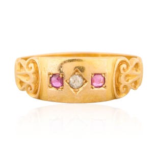 Może przedstawiać: Złoty pierścionek z prostokątną obrączką, z centralną oprawą w kształcie rombu z małym, okrągłym diamentem. Po bokach diamentu znajdują się dwie kwadratowe oprawy z różowymi kamieniami szlachetnymi. Pierścionek ma ozdobne, przypominające zwoje wzory po bokach.