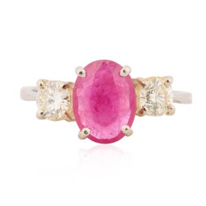 May include: A three-stone ring featuring a large oval ruby center stone flanked by two smaller, square-cut diamonds. The ring has a silver band with gold prongs holding the gemstones. The ruby is a vibrant pink color.