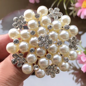 Może przedstawiać: Okrągła broszka z pereł z klastrem sztucznych pereł i błyszczącymi akcentami z cyrkonii. Broszka posiada małe, kwiatowe zdobienia z przezroczystymi cyrkoniami. Perły są kremowobiałe, a cyrkonie są przezroczyste.