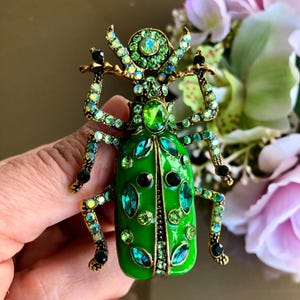 May include: A green jeweled beetle brooch with gold accents and black accents. The beetle is made of enamel and has a detailed design.