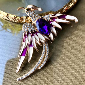 May include: A gold-toned phoenix brooch with a large purple gemstone at its center. The brooch features white and purple enamel wings, accented with clear crystals. The bird's body and tail are also embellished with crystals.