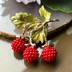 May include: A gold-toned metal brooch featuring three bright red, textured berries hanging from a gold branch with green leaves. The berries have a detailed, bumpy surface. The leaves are a vibrant green with gold accents.