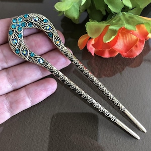 Op de afbeelding: Een paar gouden haarstokken met een decoratief ontwerp. De bovenkant van een stok heeft een bloemmotief met turquoise steentjes.