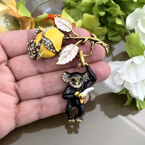 Peut inclure: Broche dorée représentant une rose jaune ornée de cristaux et un singe noir tenant une banane. Le singe et la rose sont attachés à une branche dorée avec des feuilles blanches et dorées.