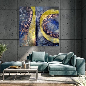 May include: Abstract painting with blue, gold, and copper tones. The painting features a large, circular shape with a smaller, circular shape inside. The painting is hanging on a gray wall in a modern living room.