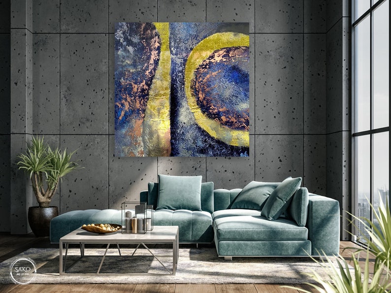 May include: Abstract painting with blue, gold, and copper tones. The painting features a large, circular shape with a smaller, circular shape inside. The painting is hanging on a gray wall in a modern living room.