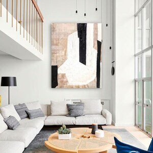 May include: A white, black, and beige abstract painting hangs on a white wall above a white sectional sofa with gray pillows. A round wooden coffee table with a woven top sits in front of the sofa. A blue armchair sits in the lower right corner of the image.