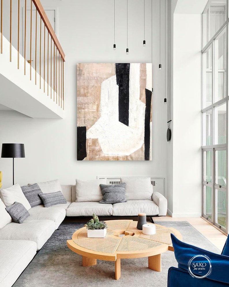 May include: A white, black, and beige abstract painting hangs on a white wall above a white sectional sofa with gray pillows. A round wooden coffee table with a woven top sits in front of the sofa. A blue armchair sits in the lower right corner of the image.