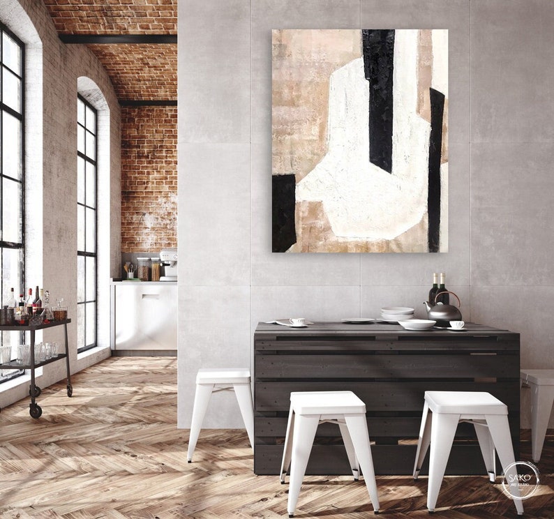 May include: Abstract painting with black, white, and beige geometric shapes on a canvas. The painting is hanging on a wall in a modern interior with a wooden table and white stools.