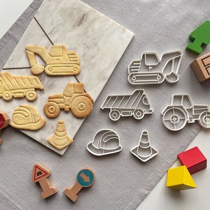 May include: A set of cookie cutters in the shape of construction vehicles, traffic signs, and trees. The cutters are made of white plastic and are arranged on a marble surface. The cutters are perfect for making cookies for a construction-themed party.