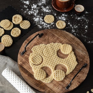 May include: A wooden chopping board with a rolling pin and a sheet of dough with round cookie cutouts. The dough has a waffle pattern and the cookies are arranged in a circle on the board.