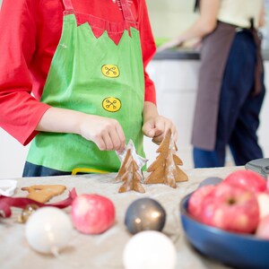 May include: A child wearing a red shirt and green apron decorates gingerbread cookies shaped like Christmas trees. The cookies are on a table with red apples, white and gray ornaments, and a brown gingerbread cookie.