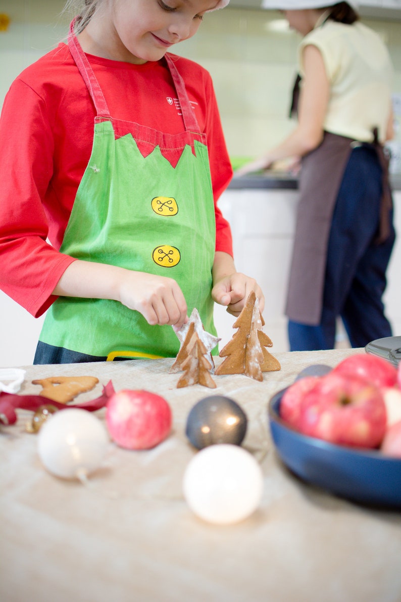 May include: A child wearing a red shirt and green apron decorates gingerbread cookies shaped like Christmas trees. The cookies are on a table with red apples, white and gray ornaments, and a brown gingerbread cookie.