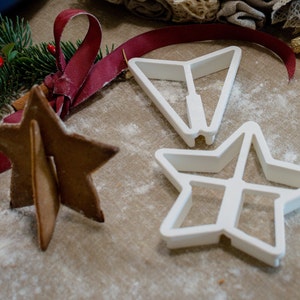 May include: Three white plastic cookie cutters in the shape of stars. The cutters are on a brown surface with white powder. A gingerbread star cookie is also on the surface.