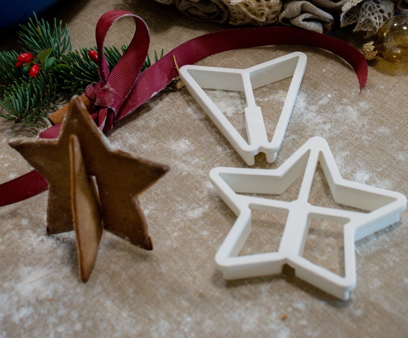 May include: Three white plastic cookie cutters in the shape of stars. The cutters are on a brown surface with white powder. A gingerbread star cookie is also on the surface.