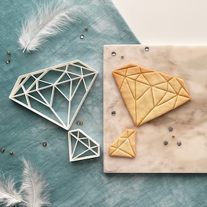 Könnte beinhalten: Drei diamantförmige Ausstecher, zwei weiße und einer beige, auf einer Marmorfläche. Die weißen Ausstecher sind geometrisch und der beige Ausstecher ist ein Keks, der mit dem Ausstecher ausgestochen wurde.