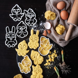 Può includere: Un set di stampini per biscotti in plastica bianca a forma di coniglietti e pulcini, alcuni dei quali sono stati utilizzati per fare biscotti di zucchero gialli su una superficie di ardesia nera. Ci sono anche due uova marroni e un mattarello su un panno di lino marrone.
