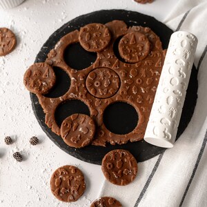 Puede incluir: Una tabla de pizarra negra con masa de galletas marrón extendida y cortada en círculos. La masa está estampada con un diseño de temática forestal. Un rodillo blanco con un diseño de temática forestal está en el lado derecho de la imagen. Hay varias galletas marrones con el mismo diseño de temática forestal en la tabla y en una superficie blanca.