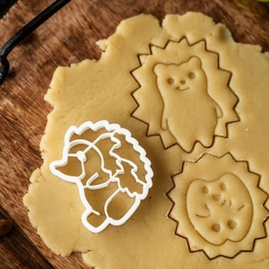 Pode incluir: Um cortador de biscoitos branco em forma de ouriço-cacheiro ao lado de um pedaço de massa com impressões de ouriço-cacheiro. A massa está numa tábua de cortar de madeira. O cortador de biscoitos é feito de plástico branco e tem um design detalhado.