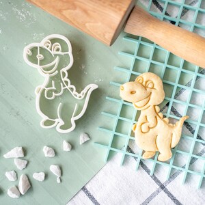 Może przedstawiać: Biała plastikowa foremka do ciasteczek w kształcie dinozaura i upieczony ciasteczko w kształcie dinozaura na niebiesko-białej powierzchni kratowej.