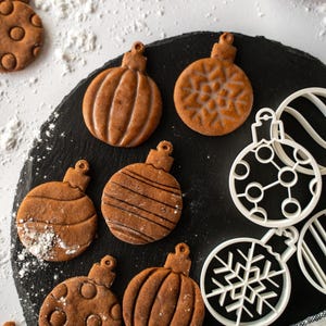 Può includere: Primo piano di biscotti di pan di zenzero a forma di decorazioni natalizie su una lavagna nera. Alcuni biscotti hanno glassa decorativa. Sono presenti anche formine bianche a forma di ornamento. Bastoncini di cannella e zucchero a velo sullo sfondo.
