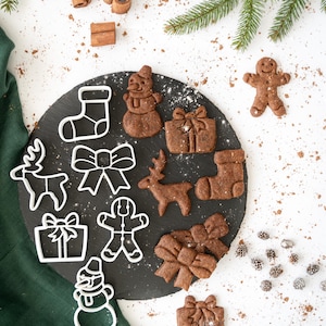 Op de afbeelding: Een set kerstkoekjesvormen op een zwarte leisteenplaat met een bestuiving van poedersuiker. De vormen zijn in de vorm van een sneeuwpop, rendier, sok, strik, peperkoekman en een cadeau. Er zijn ook een aantal peperkoekjes op de plaat en nog een paar op het witte oppervlak.