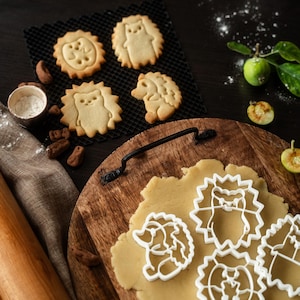 Pode incluir: Uma tábua de corte de madeira com massa de biscoito e cortadores de biscoito em forma de ouriço. Biscoitos de ouriço assados ​​estão em uma grade de resfriamento preta. Um rolo e uma tigela pequena com farinha também estão presentes.