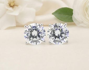May include: A pair of round-cut clear stud earrings featuring a brilliant faceted design. Each stone is secured in a four-prong silver-toned metal setting, creating a classic look with reflective surfaces that catch the light from multiple angles.