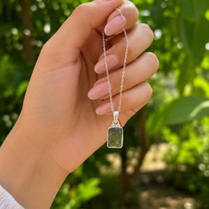 May include: A silver necklace featuring a rectangular, textured, dark green stone pendant. The pendant is held by a simple silver bail attached to a delicate silver chain. The stone has a rough, natural surface with deep green hues.