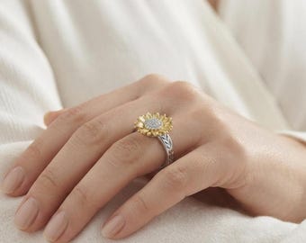 Könnte beinhalten: Ein Ring mit einem Sonnenblumenmotiv, das goldfarbene Blütenblätter und eine Mitte besitzt, auf einer silberfarbenen Ringschiene. Die Mitte der Sonnenblume ist mit kleinen, erhabenen Punkten texturiert, und die Schiene zeigt ein eingraviertes Blattmuster.