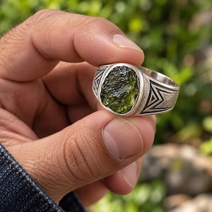 May include: A silver ring featuring a rough, textured, dark olive-green stone set in the center. The band is crafted from oxidized silver and displays a geometric pattern of etched triangles along the sides, providing a contrast to the stone.