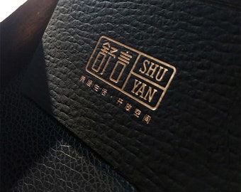 May include: A black leather business card with gold lettering. The text reads "Shu Yan" in English and Chinese characters. The card has a textured surface.