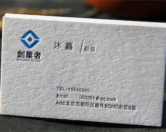 Könnte beinhalten: Ein Stapel weißer Visitenkarten mit geprägtem blauem und schwarzem Text. Die Karten zeigen ein Logo und chinesische Schriftzeichen sowie Kontaktinformationen wie Telefonnummer, E-Mail und Adresse.