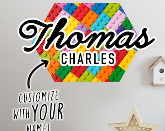 Puede incluir: Una calcomanía de pared hexagonal de colores con un diseño similar al Lego. La calcomanía presenta el nombre "Thomas Charles" en una fuente cursiva negra. Una flecha apunta al nombre, con el texto "¡Personaliza con TU NOMBRE!" debajo.