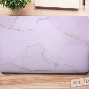 Puede incluir: Una piel para portátil de color lila claro con un patrón de mármol y detalles dorados. La piel está diseñada para adaptarse a un portátil y tiene un acabado liso y mate. La piel se muestra sobre una superficie de madera con otros artículos de oficina.