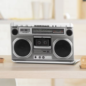 May include: A silver and black vintage boombox with a cassette player and radio tuner. The boombox has two large speakers and a variety of buttons and knobs.