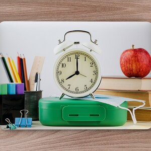 May include: A white alarm clock set to 7:00, a green lunchbox, a red apple, and a stack of books on a wooden surface. A face mask is on top of the books. There are also pencils, a ruler, and a pen holder with a pen and a compass in it.
