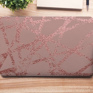 Peut inclure: Un ordinateur portable avec un motif géométrique rose gold sur le couvercle. L'ordinateur portable est sur une surface en bois avec d'autres fournitures de bureau.