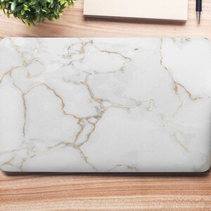 May include: A white laptop case with a marble pattern and gold veins. The case is on a wooden surface with other office supplies.